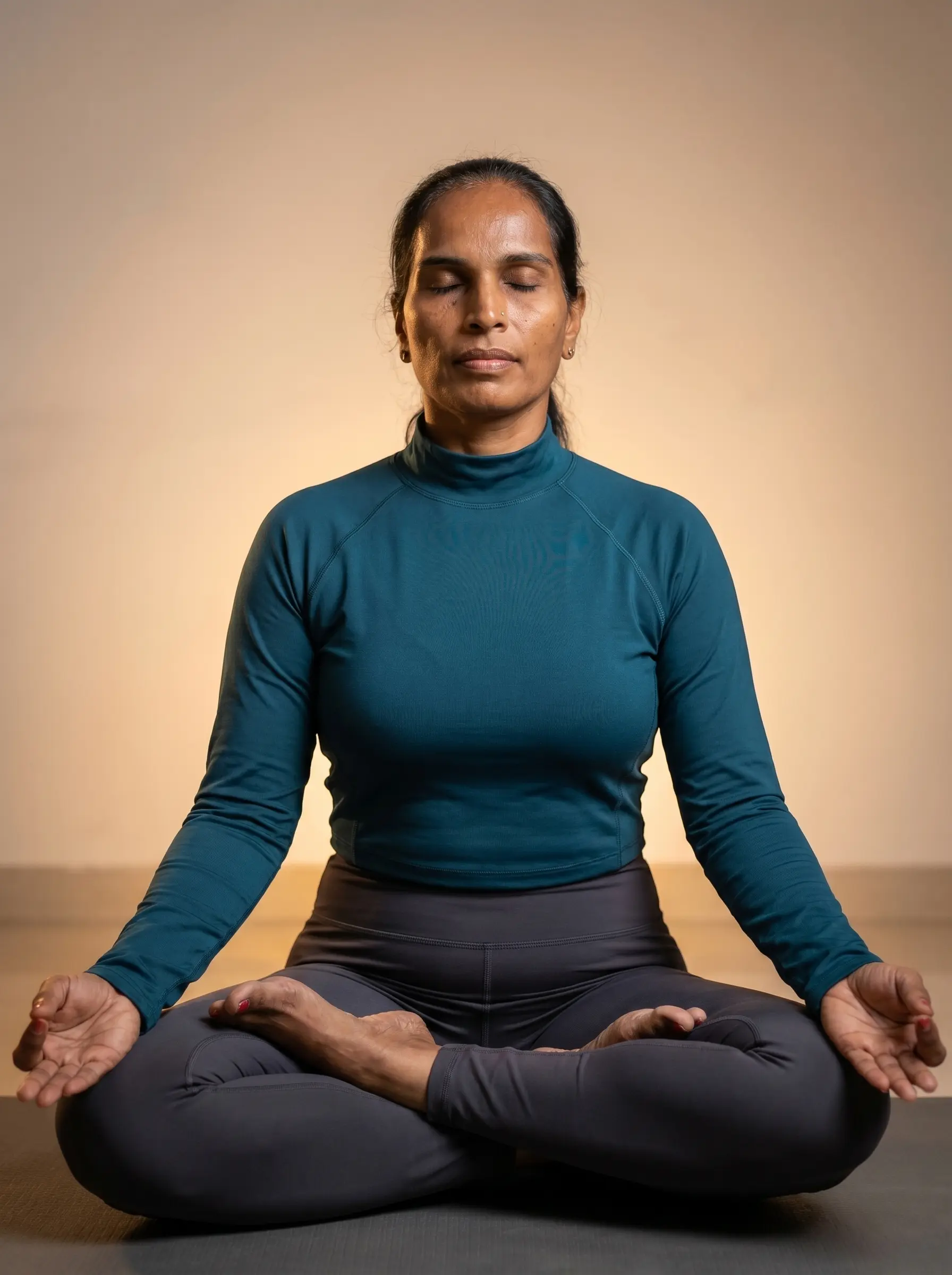 A peaceful Geetanjali with eyes closed in Lotus pose for Pranayama and Meditation, wearing her signature deep teal athletic top.